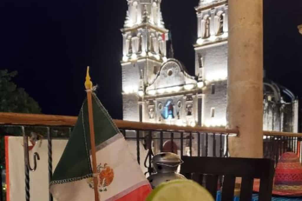 This image beautifully frames a nighttime view of the illuminated Campeche Cathedral in Mexico. In the foreground, a margarita garnished with lime sits on a table alongside a small Mexican flag, creating a festive and cultural ambiance. The warmly lit historic architecture contrasts against the dark night sky, enhancing the charm of this dining setting.