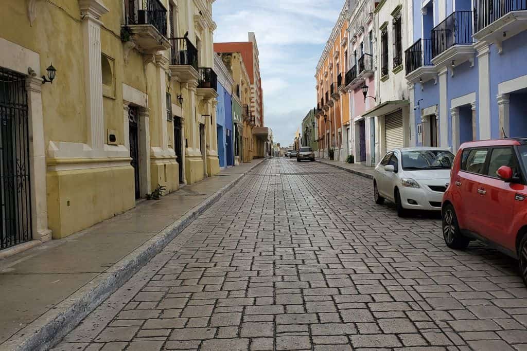 A cobblestone street in Campeche lined with colorful colonial-style buildings stretches into the distance. Parked cars sit along the narrow road, and balconies overlook the quiet scene.