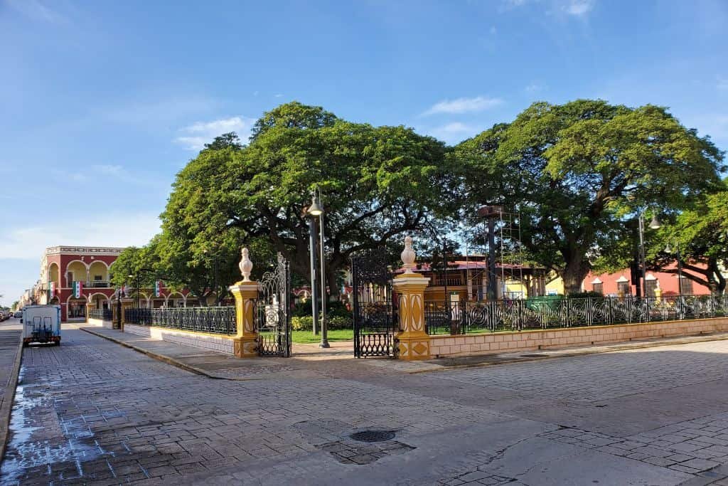 This image captures a scenic town square in Campeche, Mexico, featuring lush green trees and a decorative wrought-iron fence. The golden-hued entrance pillars add a touch of colonial charm, complementing the colorful historic buildings in the background. The cobblestone streets and classic street lamps enhance the picturesque, old-world atmosphere.
