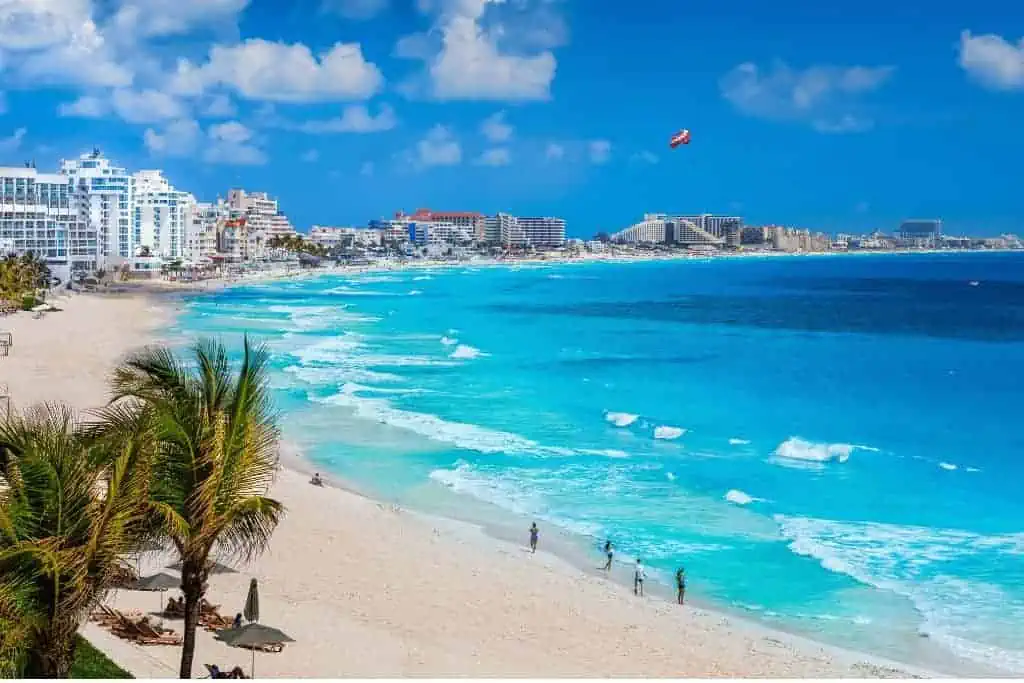 A beach in Cancun, Mexico, featuring turquoise blue waters gently crashing onto the sandy shoreline. In the background, a row of modern beachfront hotels lines the coast, while palm trees and a few people enjoying the beach are visible in the foreground, set against a bright blue sky with scattered clouds.