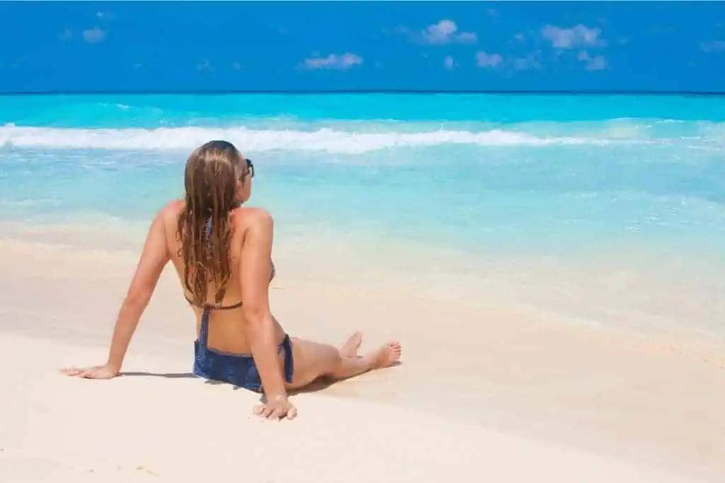 A woman sitting on a sandy beach, facing the bright turquoise ocean under a clear blue sky. She is wearing a bikini and sunglasses, relaxing with her hands resting on the sand as she enjoys the peaceful, tropical surroundings.