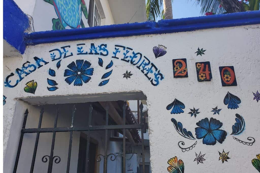 The image shows a white wall with hand-painted floral designs in blue and green, surrounding the words “Casa de las Flores.” The house number “240” is displayed in red and black tiles. A black metal gate is visible beneath the artwork, leading into the property.