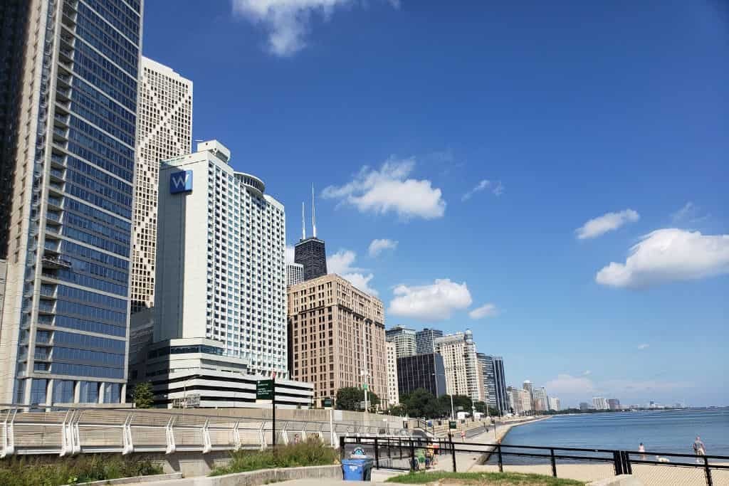 Tall skyscrapers line a waterfront promenade with a clear view of the lake and blue sky. Pedestrians walk along the path bordered by railings and greenery.