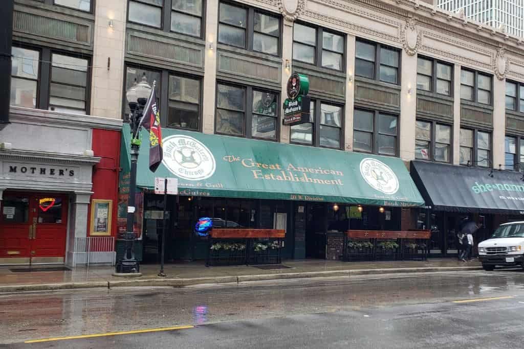 The image shows a street view of an Irish pub with a large green awning reading “The Clover Irish Pub and Restaurant.” The building has tall windows and decorative stonework, with rainbow and Irish flags hanging outside. The sidewalk and street are wet from rain, and a white vehicle is parked nearby.