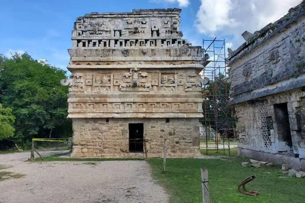A detailed view of a stone structure from the ancient Mayan ruins of Chichen Itza. The building features intricate carvings and stone patterns, with various geometric shapes and symbols typical of Mayan architecture. Surrounding the structure are grassy areas, trees, and scaffolding on the right side, indicating ongoing restoration or preservation work.