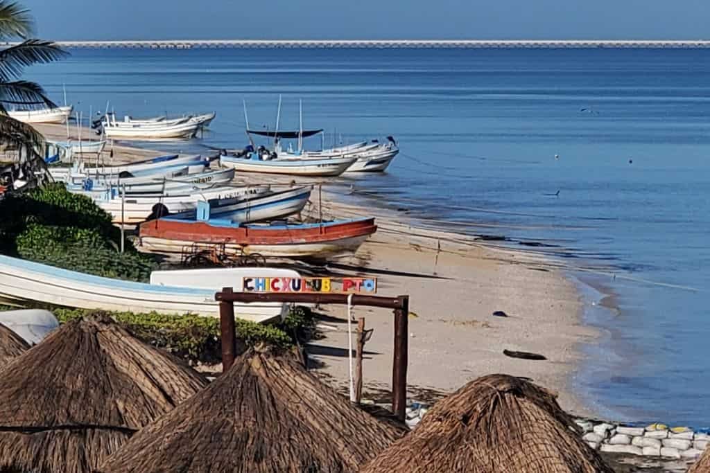 This image showcases a serene beach scene with small fishing boats lined up along the sandy shore in Chicxulub Puerto, Mexico. Thatched-roof palapas are visible in the foreground, adding a rustic, tropical charm. The calm blue waters stretch toward the horizon, creating a peaceful coastal atmosphere.