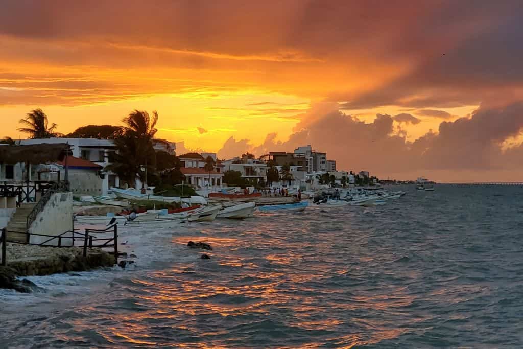 A vibrant sunset casts golden and orange hues across the sky above a coastal town, with waves gently crashing against the shore. Palm trees, houses, and small boats line the waterfront, creating a peaceful seaside scene in Chicxulub Puerto, Mexico.