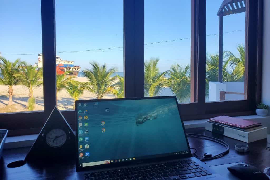 This image showcases a scenic workspace setup with a laptop on a wooden desk facing a large window. The view outside features palm trees, a sandy beach, and a colorful boat under a bright blue sky. The serene coastal setting creates an ideal environment for remote work or relaxation.