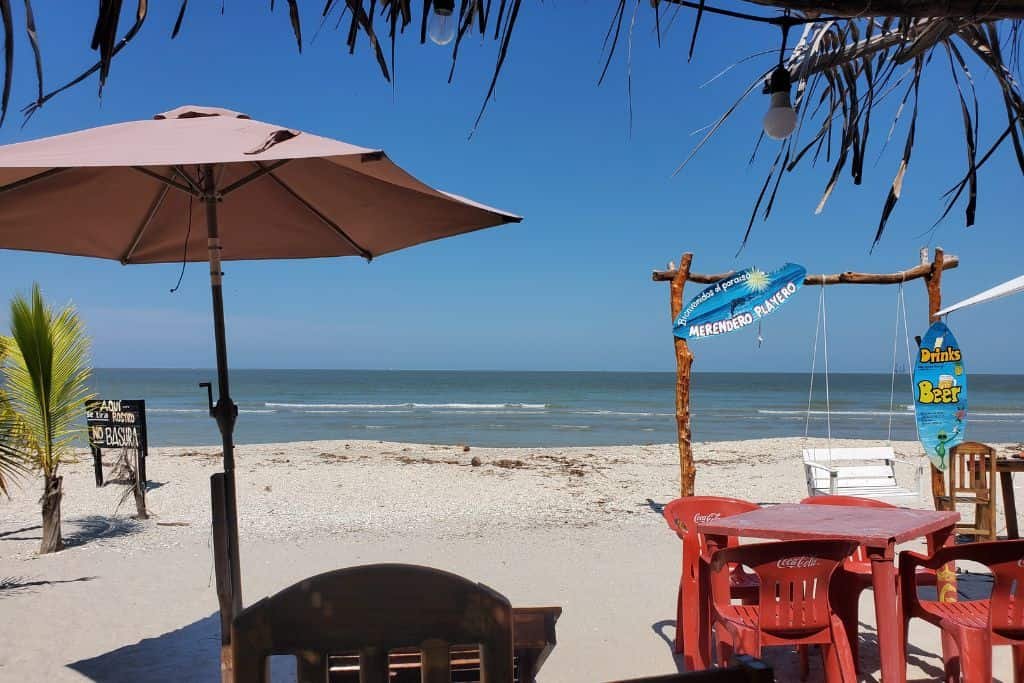 This image captures a relaxing beachfront restaurant scene in Ciudad del Carmen, Mexico. A shaded table with red chairs sits on the sandy shore, overlooking calm ocean waves. A wooden swing adorned with beach-themed signs adds to the laid-back, tropical vibe.