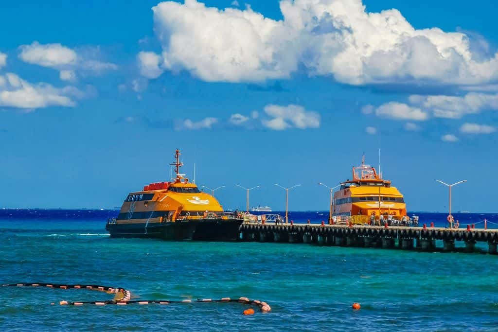 Two bright yellow Ultramar ferries are docked at a long pier extending into turquoise water. The vibrant scene is set under a blue sky with scattered white clouds.