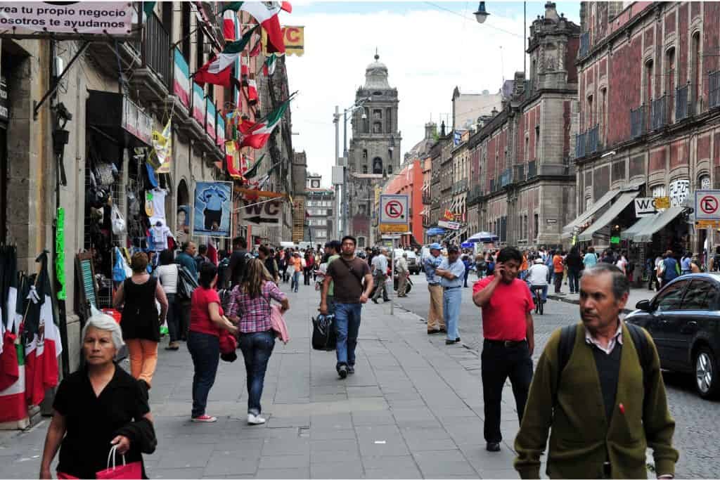 A busy street in Mexico City with many people walking on the sidewalk. The buildings are old and beautiful. One building has many flags in front of it.