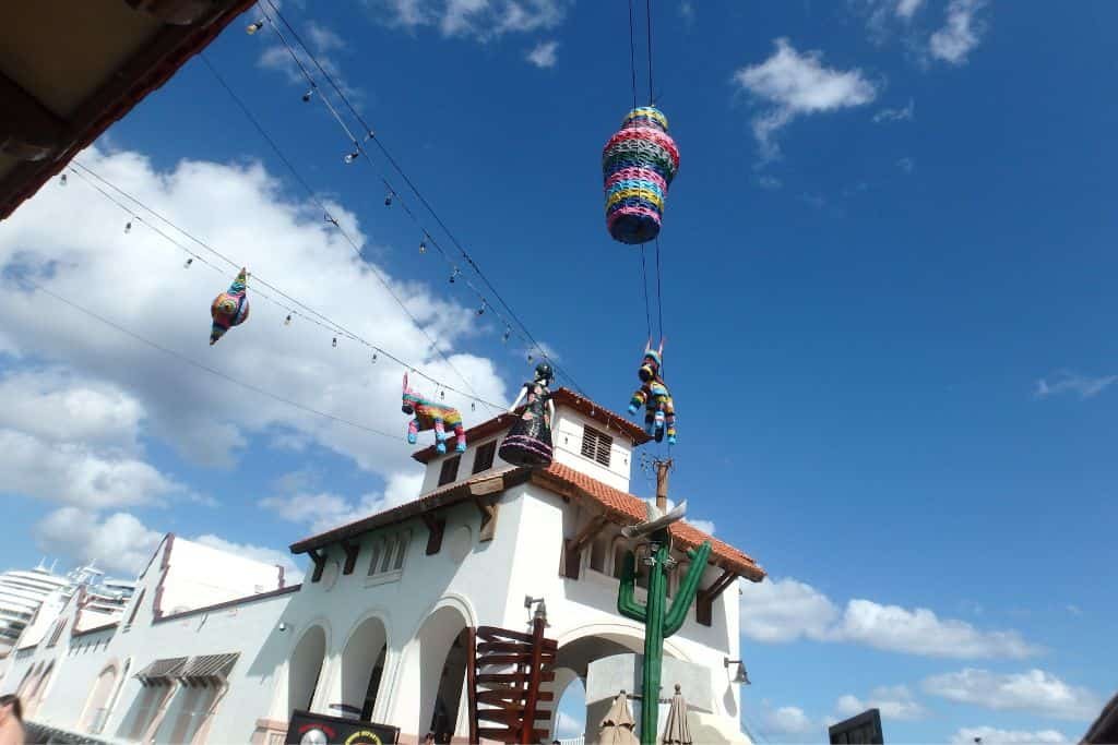 This image features a bright blue sky with scattered clouds above a white building with arched windows and a red-tiled roof. Several piñatas, including a large, colorful cylindrical one, hang from wires stretching across the scene.