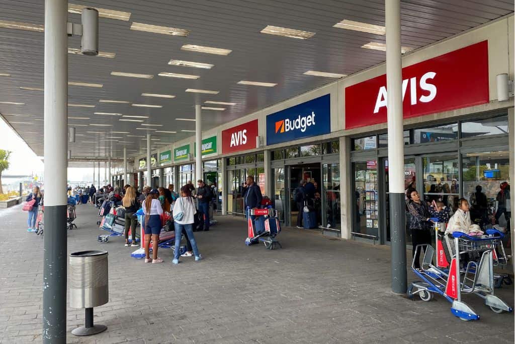 Travelers queue at rental car service desks for Avis and Budget at an airport.