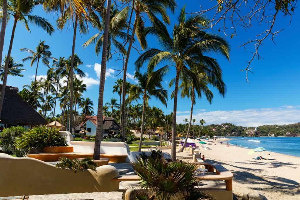 This image shows a tropical beach lined with tall palm trees and bright blue skies. People are relaxing on the sandy shore, with beachside villas and greenery adding to the resort-like atmosphere.