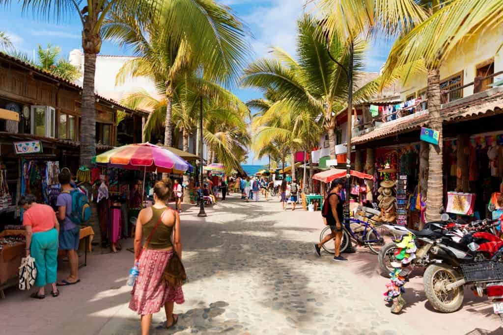 A lively pedestrian street lined with palm trees features colorful market stalls and shops. People stroll with umbrellas and bicycles, creating a vibrant tropical shopping scene.