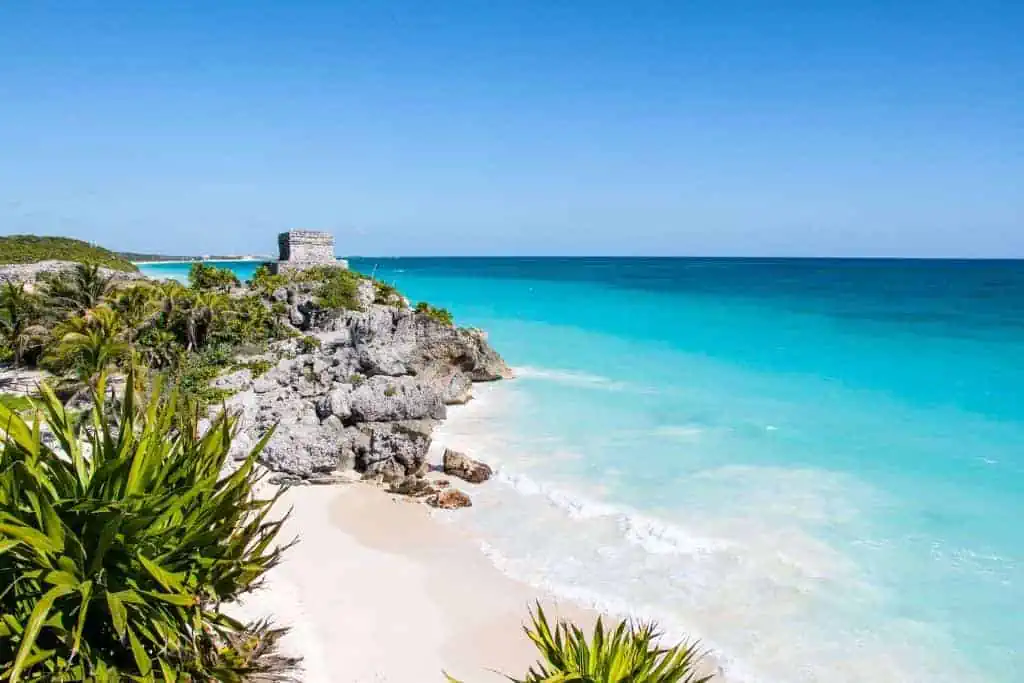 The stunning coastline of Tulum, with ancient Mayan ruins sitting atop a rocky cliff overlooking the turquoise ocean. The white sandy beach is framed by lush green vegetation, and the calm sea contrasts beautifully with the clear, blue sky above.