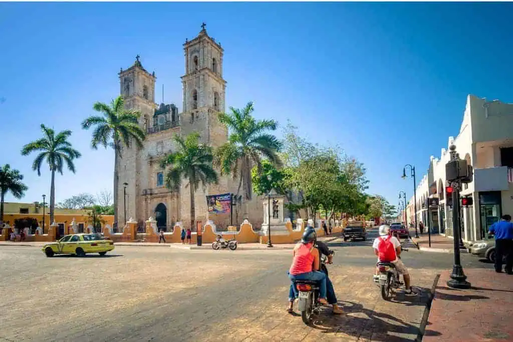 A town square in Valladolid, Mexico, with a historic church as the central focus, characterized by its twin towers and classic colonial architecture. Palm trees and people on motorcycles add to the lively street scene, while a bright blue sky serves as the backdrop, highlighting the charm and cultural atmosphere of the town. A yellow taxi is seen driving through the square.