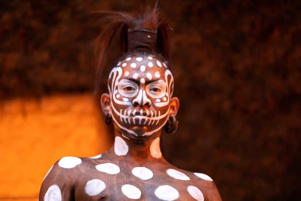 A performer at Xcaret Park in Mexico, dressed in traditional Mayan ceremonial body paint. His face and upper body are covered in striking white patterns on a dark base, resembling a skull and polka dots. He has an intense gaze, tribal earrings, and a distinctive hairstyle, set against a dimly lit, earthy-toned background.