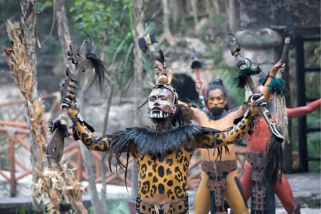 Performers at Xcaret reenact an ancient Mayan ceremony, dressed in elaborate costumes with face paint and animal-inspired designs. The central figure wears a jaguar-patterned outfit with black feathered accents, holding ceremonial staffs adorned with hair and bones. Behind him, other performers in tribal attire raise their arms in a dramatic ritual setting, surrounded by stone structures and lush vegetation.