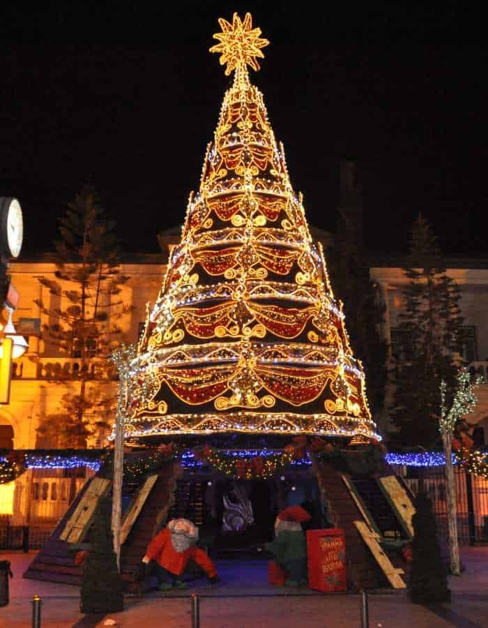 Christmas in Limassol features a grand, cone-shaped Christmas tree adorned with golden lights, red ornaments, and garlands, topped with a radiant star. At the base, playful holiday figures and gift boxes create a cheerful, family-friendly holiday scene.
