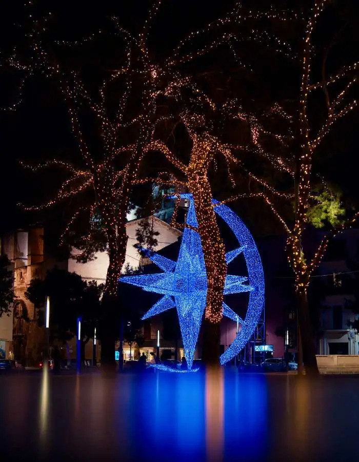Christmas in Palma de Mallorca is marked by a glowing blue sculpture of a star and crescent moon set among trees wrapped in golden string lights. The festive installation is reflected on a smooth surface below, creating a dreamy nighttime holiday scene.