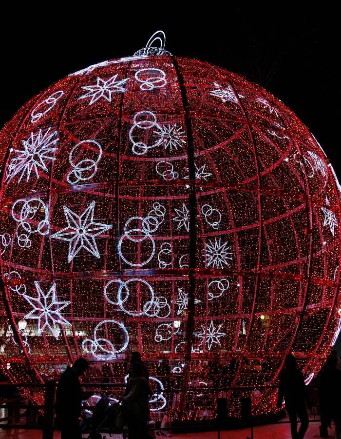 Christmas in Alicante features a massive red and white illuminated ornament structure decorated with stars and circles. Silhouettes of people walking beneath it highlight its grand scale and create a magical, immersive holiday experience.