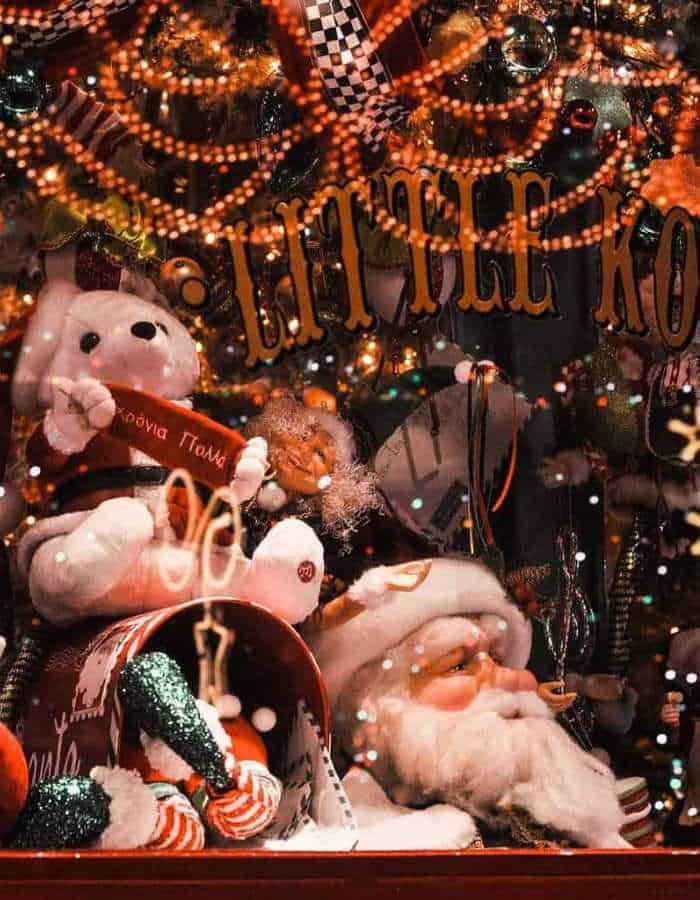 A festive shop window display for Christmas in Athens features a plush polar bear, Santa Claus figure, candy canes, and twinkling ornaments under the sign “Little Ko.” The scene is bursting with holiday cheer, showcasing traditional Christmas colors and decorations.