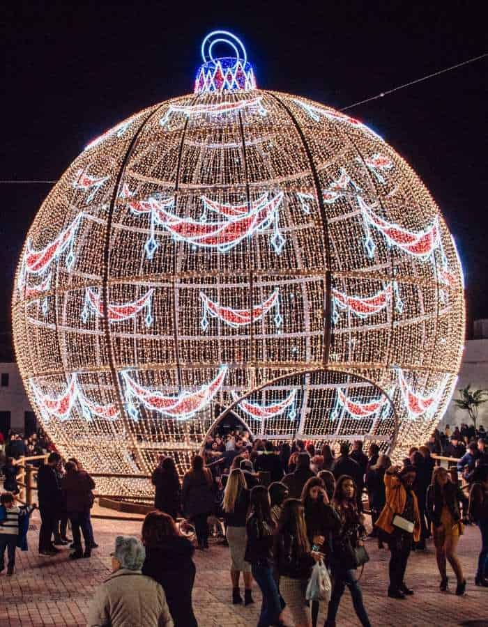 Christmas in Cádiz comes alive with a massive illuminated ornament structure glowing in warm white, red, and blue lights. Crowds of people gather around and walk through the festive centerpiece, adding energy to the holiday night scene.