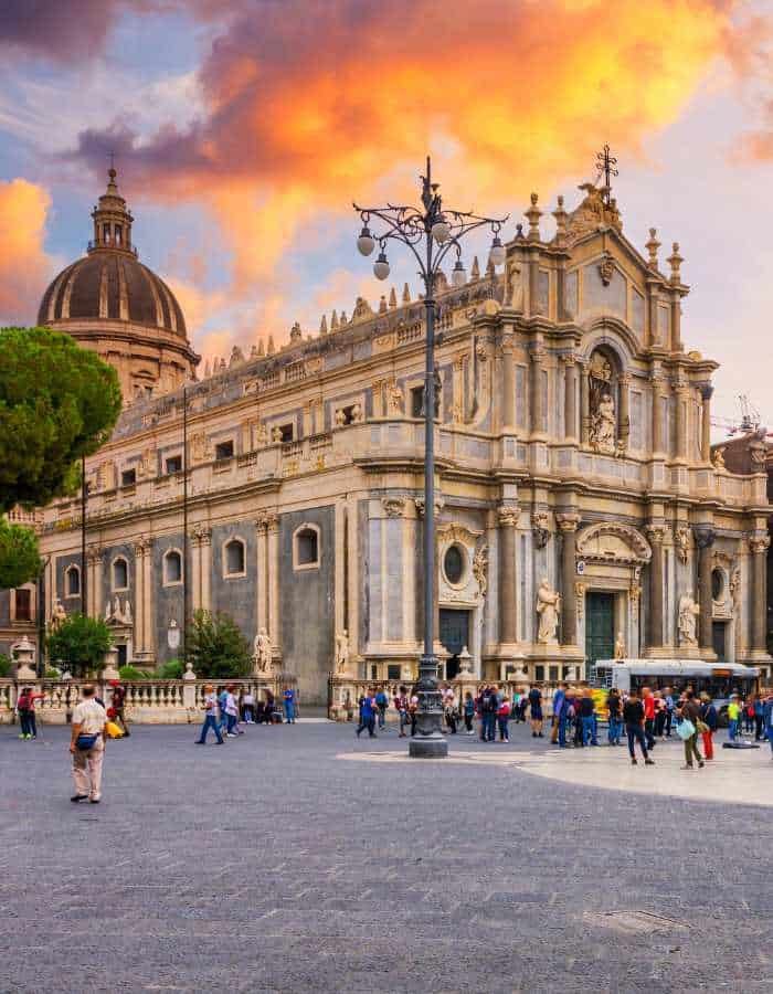 Christmas in Catania unfolds in front of the stunning Catania Cathedral, where crowds gather in the square under a fiery orange sunset sky. The grand baroque architecture sets a majestic backdrop for the city's festive atmosphere.