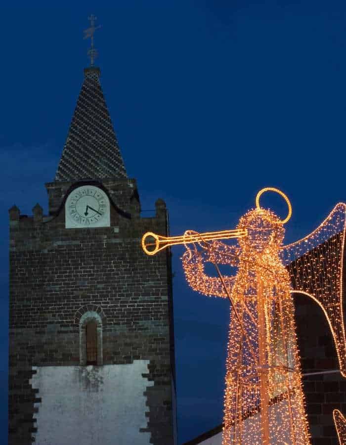 Christmas in Funchal features a glowing angel made of golden lights blowing a trumpet in front of a historic clock tower. The deep blue evening sky adds contrast to the warm illumination, highlighting the festive spirit in the city’s old town.
