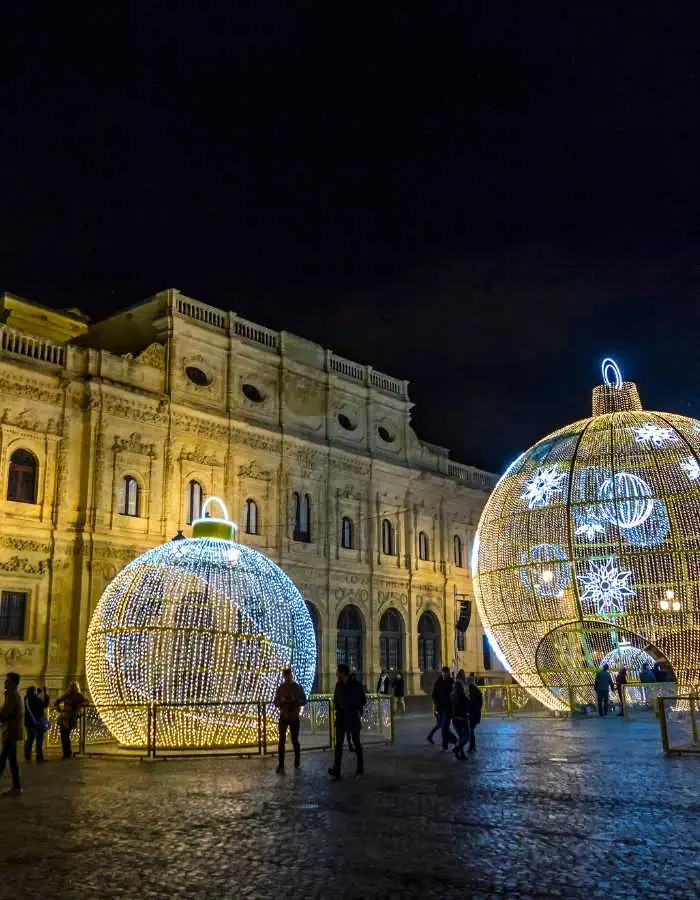 Two giant, glowing Christmas ornaments lit with white and golden lights sit in front of a historic building during Christmas in Seville. People walk around and through the illuminated structures, adding a sense of wonder to the festive evening scene.