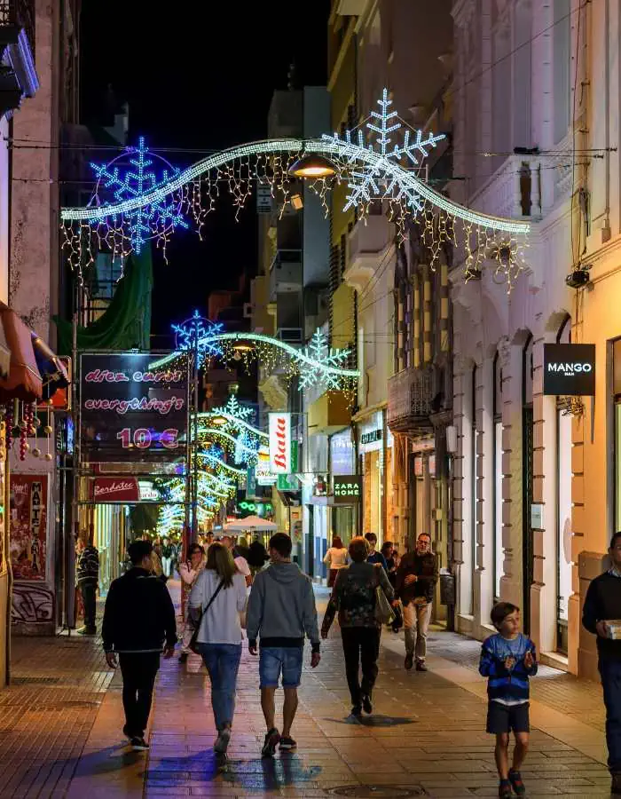 Christmas in Tenerife is celebrated with glowing snowflake lights strung above a busy shopping street lined with stores and holiday shoppers. The festive decorations add a wintery charm to the mild island evening.