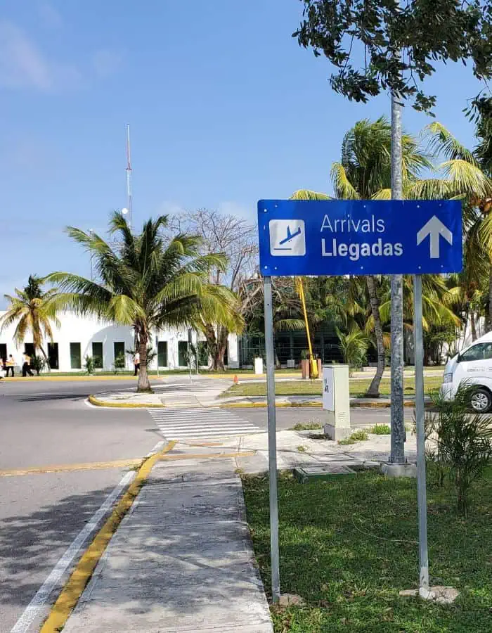 A blue bilingual sign reading “Arrivals / Llegadas” with an arrow points toward the arrivals area at Cozumel Airport. Palm trees and a white terminal building are visible in the background under a clear, sunny sky.