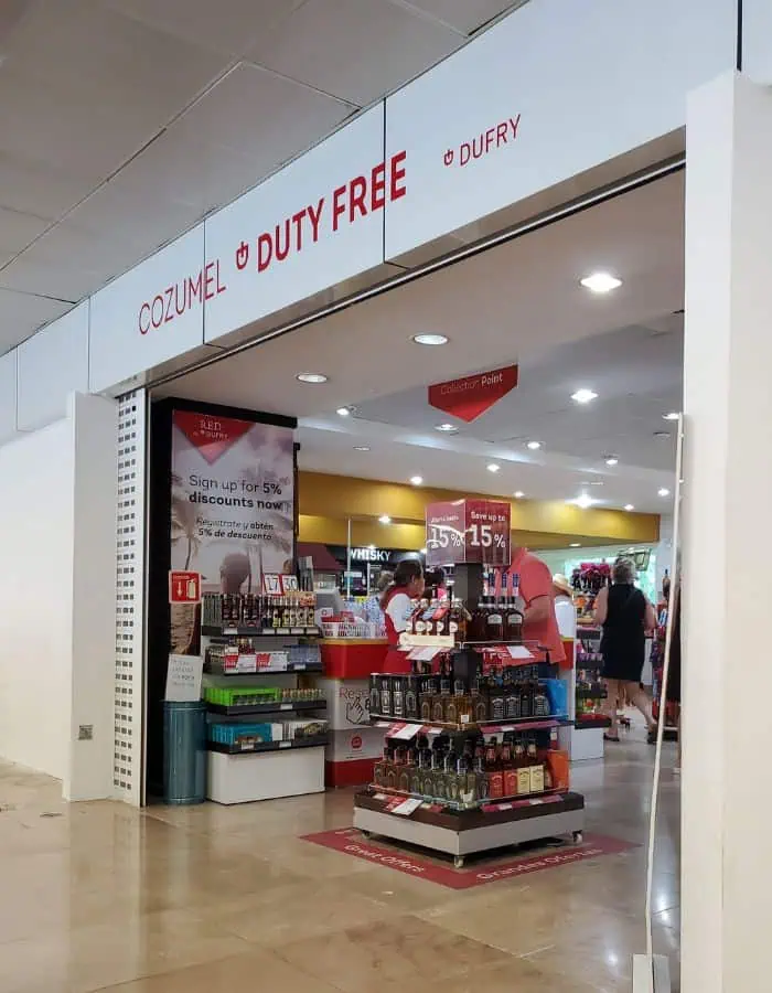 Entrance to the Cozumel Duty Free store operated by Dufry, displaying shelves of liquor, promotional signage offering up to 15% off, and a checkout counter. Travelers browse the brightly lit shop, which features signs in both English and Spanish.