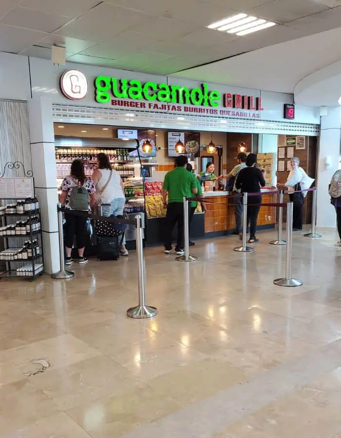 Travelers wait in line at Guacamole Grill, a casual airport restaurant in Cozumel offering burgers, fajitas, burritos, and quesadillas. The bright sign and open counter setup invite quick service, with snacks and drinks visible behind the staff preparing orders.