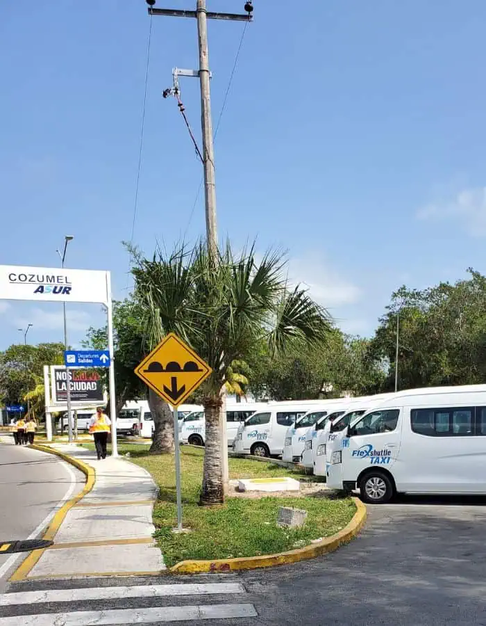 A row of white FlexShuttle Taxi vans lines the curb outside Cozumel Airport under a clear blue sky. A yellow speed bump warning sign and Cozumel ASUR signage are visible, along with palm trees and airport staff walking along the sidewalk.