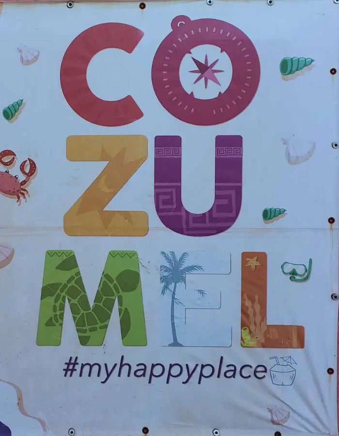 This image features a colorful sign that reads **"COZUMEL #myhappyplace"** with each letter decorated with tropical and cultural elements. The background includes playful illustrations of a crab, seashells, a turtle, and a palm tree, enhancing the island vibe. The design reflects the joy and beauty of Cozumel, making it a perfect photo spot for visitors who love this tropical paradise.