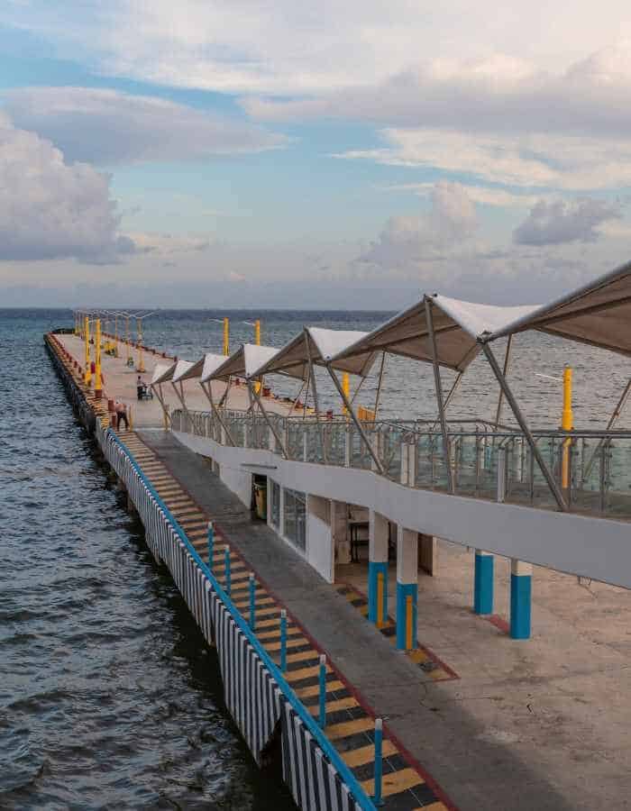 Long pier with covered seating areas extends over the water, supported by blue and yellow pillars. The scene is set under a partly cloudy sky, with calm waves along the structure’s edge.