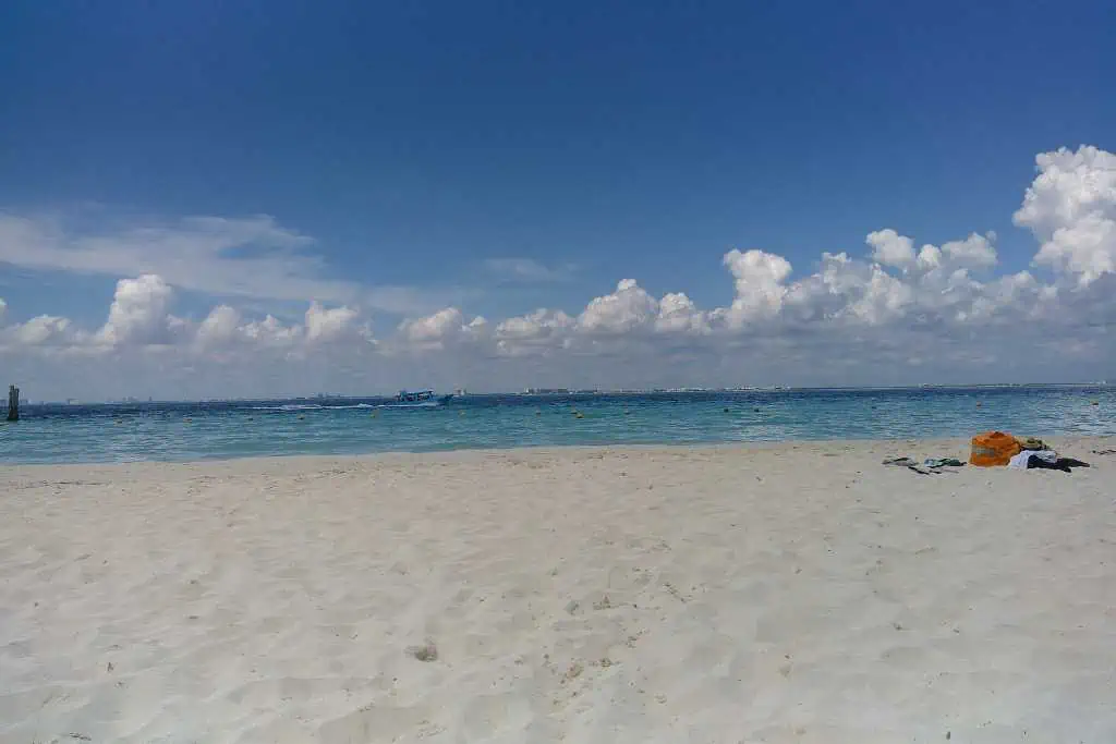 A sandy beach with white sand leading to calm blue ocean water under a bright sky with scattered clouds. A few belongings, such as towels or bags, are placed on the sand near the shoreline. The horizon is visible in the distance, giving the scene an open and tranquil feel.