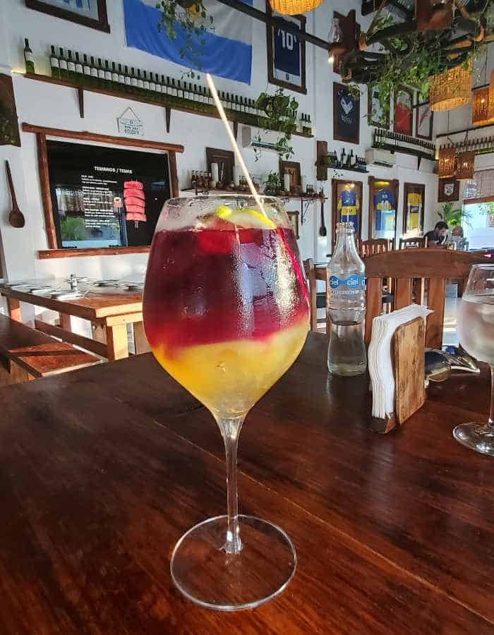 A large wine glass filled with layered sangria—deep red on top and bright yellow-orange on the bottom—sits on a wooden restaurant table in Cozumel. The background features a cozy interior with framed artwork, flags, and hanging plants, capturing a colorful and eclectic tropical vibe.