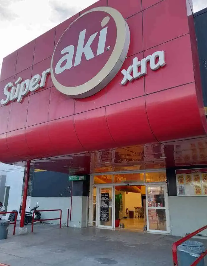 Entrance of a Súper Aki Xtra store with a bold red and white exterior and large logo above the doors. A scooter and shopping cart are near the entrance, suggesting a convenient neighborhood supermarket.