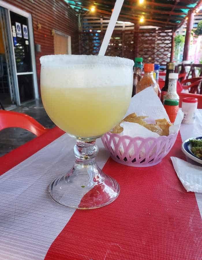 Large margarita glass filled with a frozen yellow drink sits on a red and white tablecloth. Nearby are a basket of tortilla chips, bottles of hot sauce, and a bowl of salsa in a casual open-air restaurant setting.