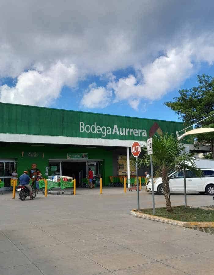 Exterior of a Bodega Aurrera store with a bright green facade and white lettering. People enter and exit the store, while a scooter and cars are parked outside under a partly cloudy sky.