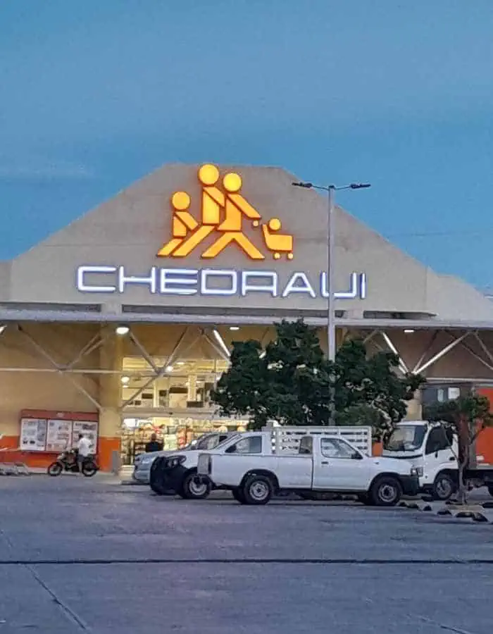 Chedraui supermarket exterior at dusk features its illuminated logo of a family with a shopping cart above the entrance. Several white trucks are parked in front of the well-lit storefront.