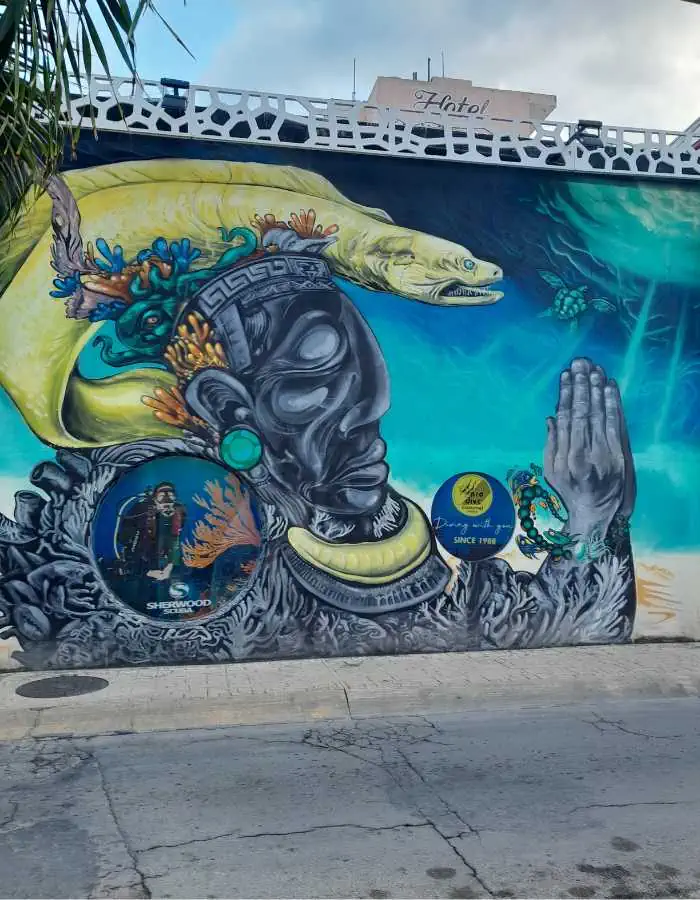A vibrant mural on a Cozumel street features a stylized figure with hands in prayer, adorned with Mayan-inspired jewelry and surrounded by ocean elements like coral, fish, and a large yellow eel. The artwork blends cultural and marine imagery, celebrating the island’s heritage and sea life.