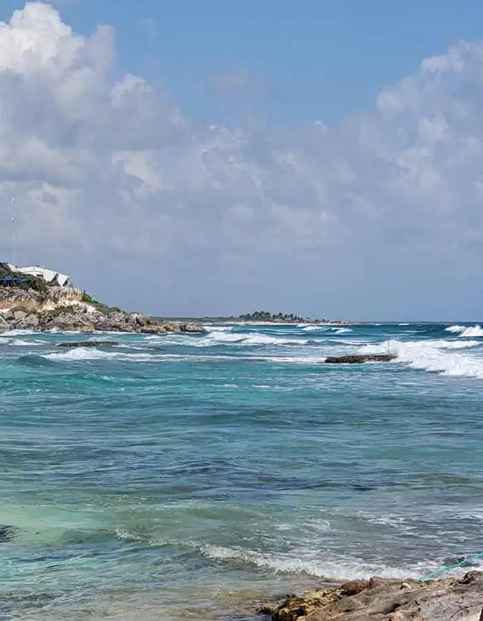Waves roll over the rocky shoreline at Playa Chen Rio in Cozumel, with clear turquoise water and a rugged coastline stretching into the distance. White clouds fill the sky, and a few buildings are perched on the cliffside.