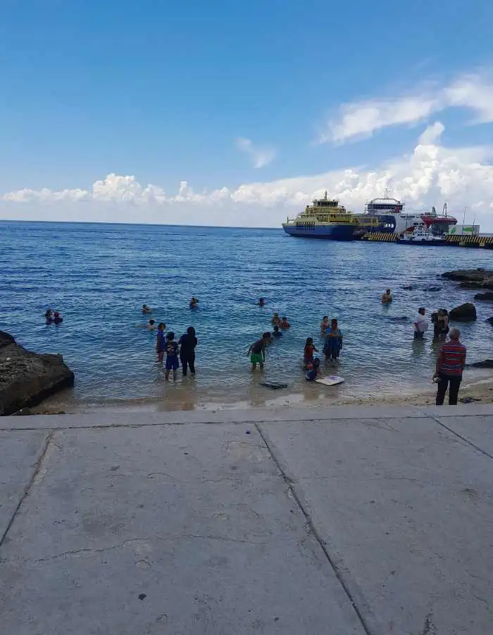 Locals and visitors swim and wade in the shallow water at Playa Caletitas in Cozumel, just off a concrete walkway. In the distance, large ferries are docked under a blue sky with puffy white clouds.
