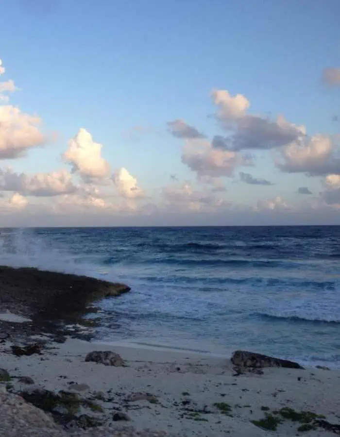 Waves crash against the rocky shoreline at Playa Corona in Cozumel as the sun sets, casting a warm glow on the clouds scattered across the sky. The beach is lined with sand, stones, and patches of greenery, with the ocean stretching into the horizon.