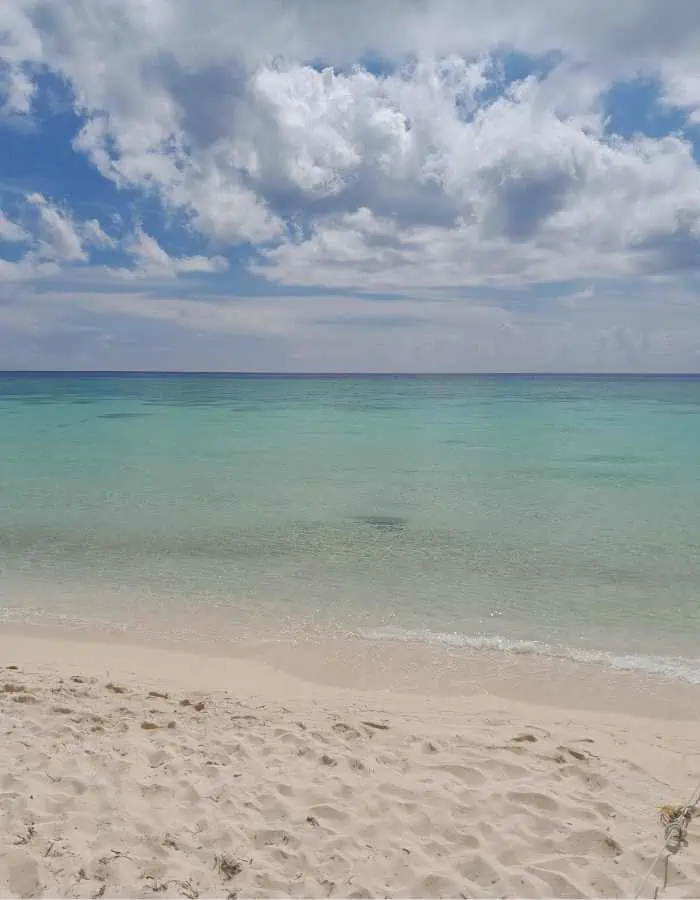 A serene view of Playa Palancar in Cozumel with powdery white sand leading to clear, shallow turquoise waters and a calm sea under a sky filled with scattered, fluffy clouds.