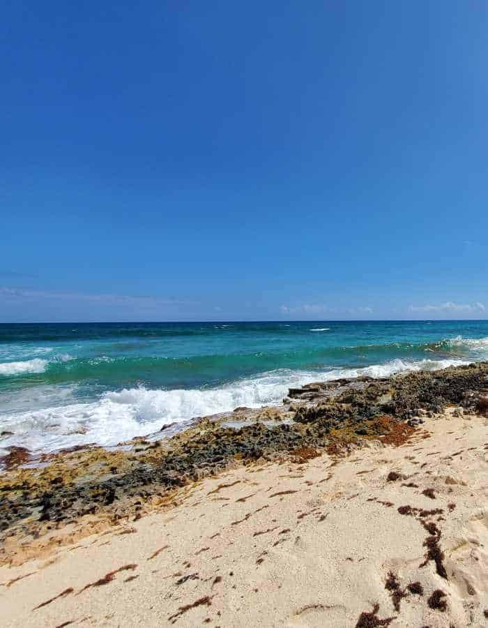 Bright, sunny day at Playa San Martín in Cozumel with soft golden sand, rugged rocky shoreline, and turquoise waves crashing gently under a cloudless blue sky.